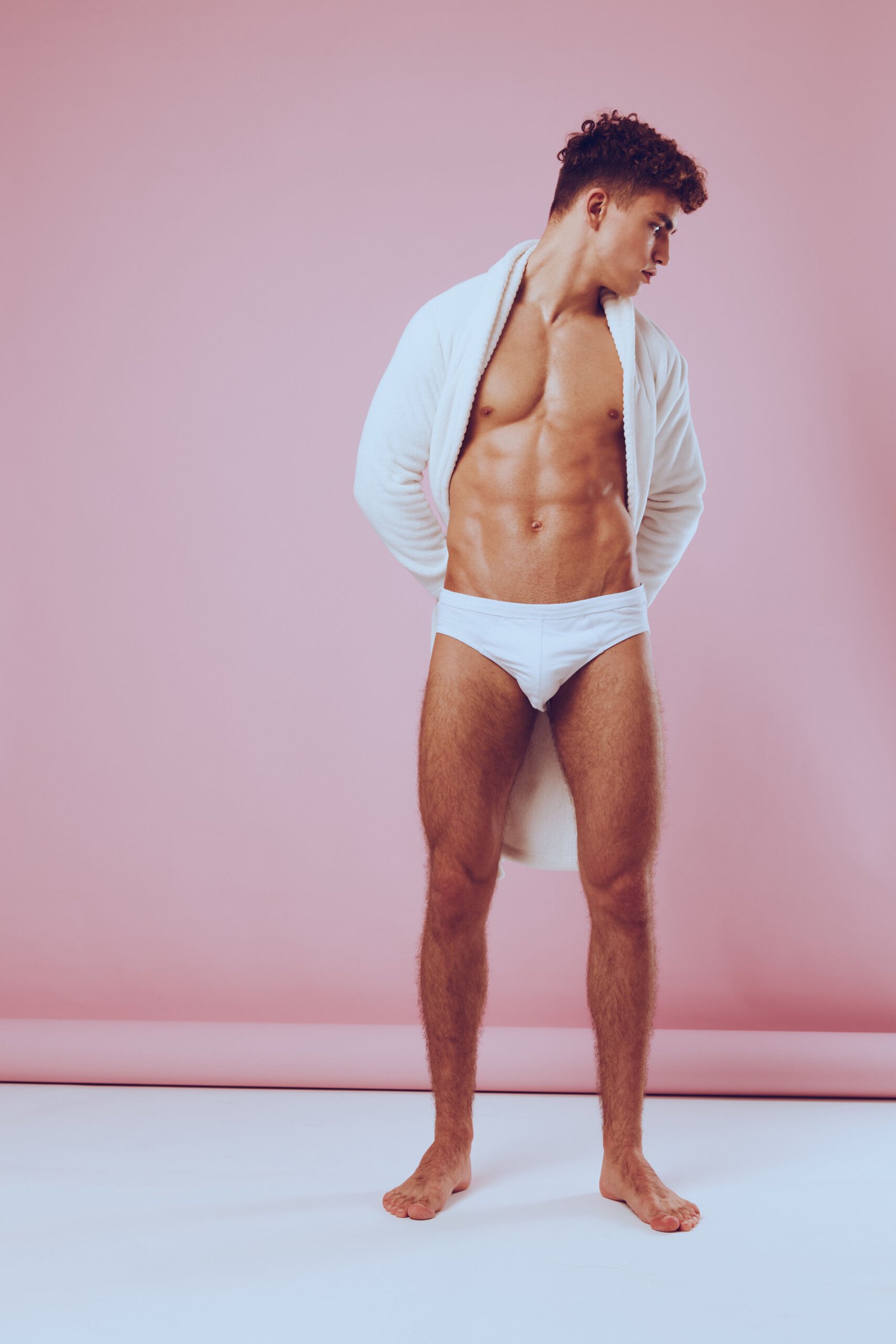 Confident young man posing in white underwear and a robe, showcasing his athletic physique against a soft pink background. Fashion and body confidence concept.